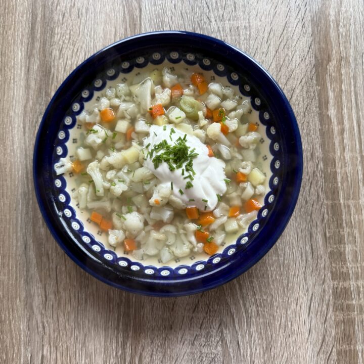 A bowl of Zupa Jarzynowa, a traditional Polish vegetable soup with diced carrots, potatoes, and herbs, is topped with a dollop of sour cream and chopped parsley, served on a wooden table.