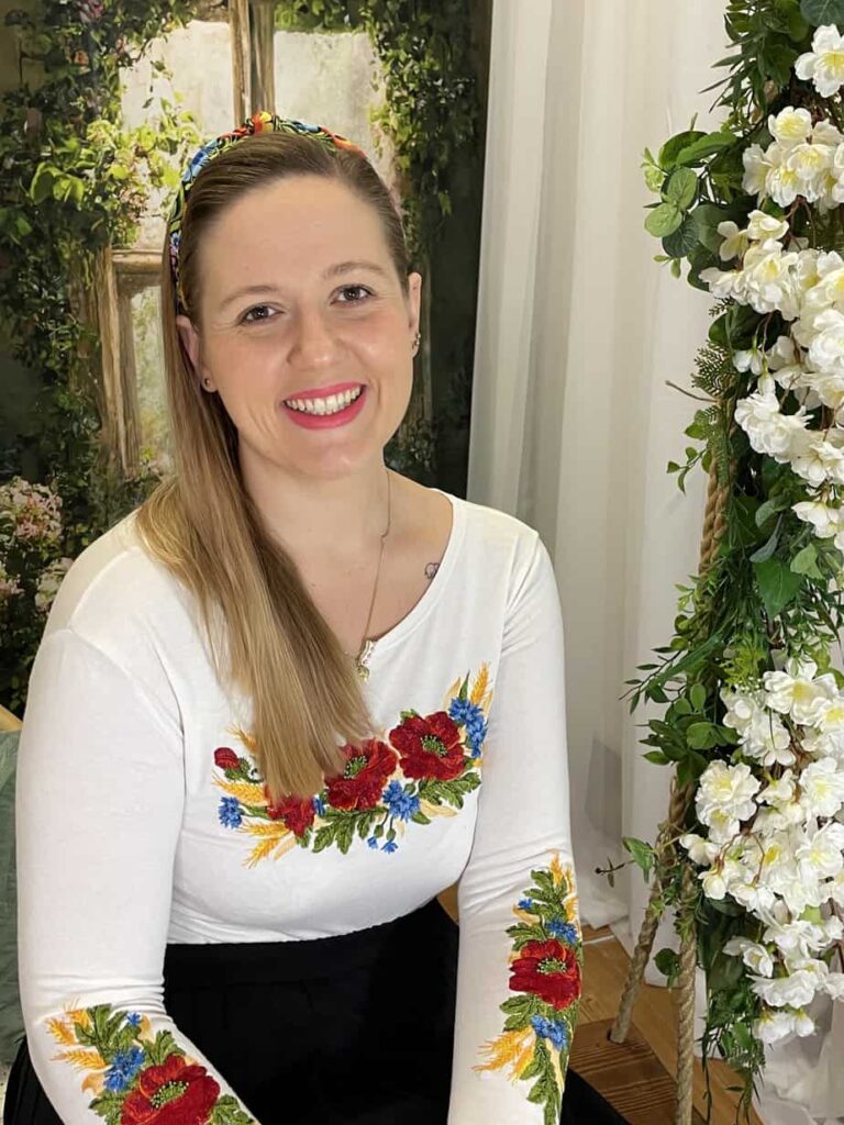 A smiling woman wearing a floral headband and a white top with vibrant flower embroidery, seated next to white flowers, becomes the captivating focus of our latest blog post.