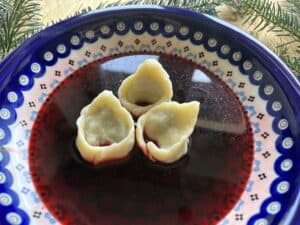 A ceramic plate with three uszka dumplings in a dark red soup, reminiscent of a traditional Polish recipe, with a decorative evergreen branch in the background.