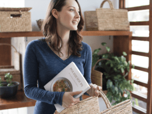 A woman holds a book titled "Polish Soups" and a wicker basket while standing in a room adorned with wicker baskets and plants, showcasing her passion for Polish recipes.
