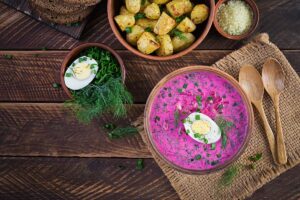 A vibrant flat lay of a traditional Polish summer food with a bowl of beetroot soup, accompanied by potatoes, chopped herbs, a boiled egg, and wooden spoons on a rustic table.