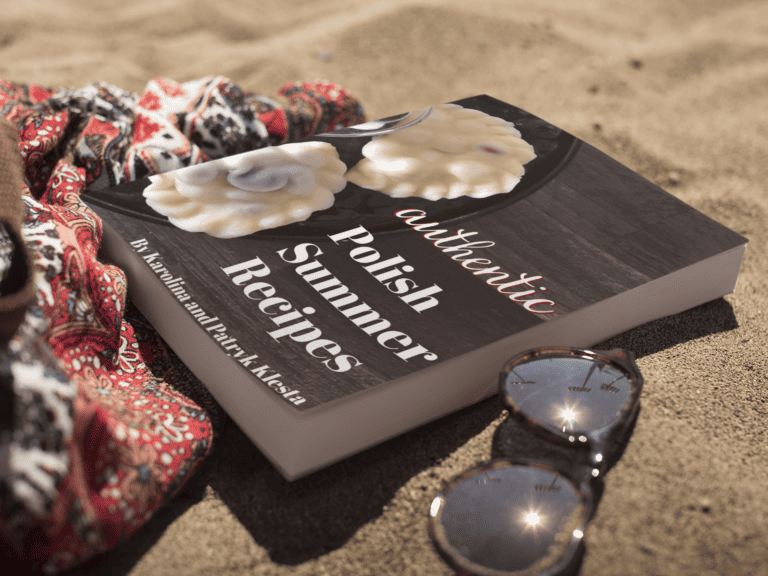A cookbook titled "Authentic Polish Summer Recipes" lies on sand next to sunglasses and a patterned cloth.