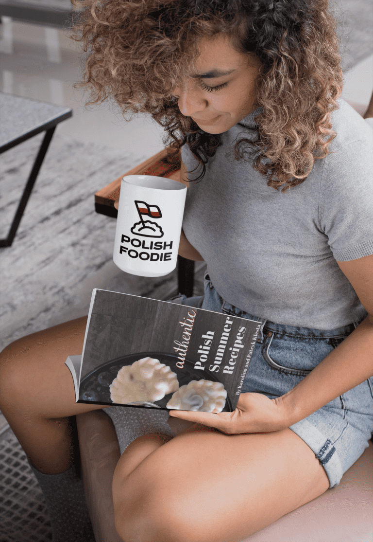 A person with curly hair sits on a sofa, holding a "Polish Foodie" mug and reading a cookbook filled with authentic Polish Summer Recipes.