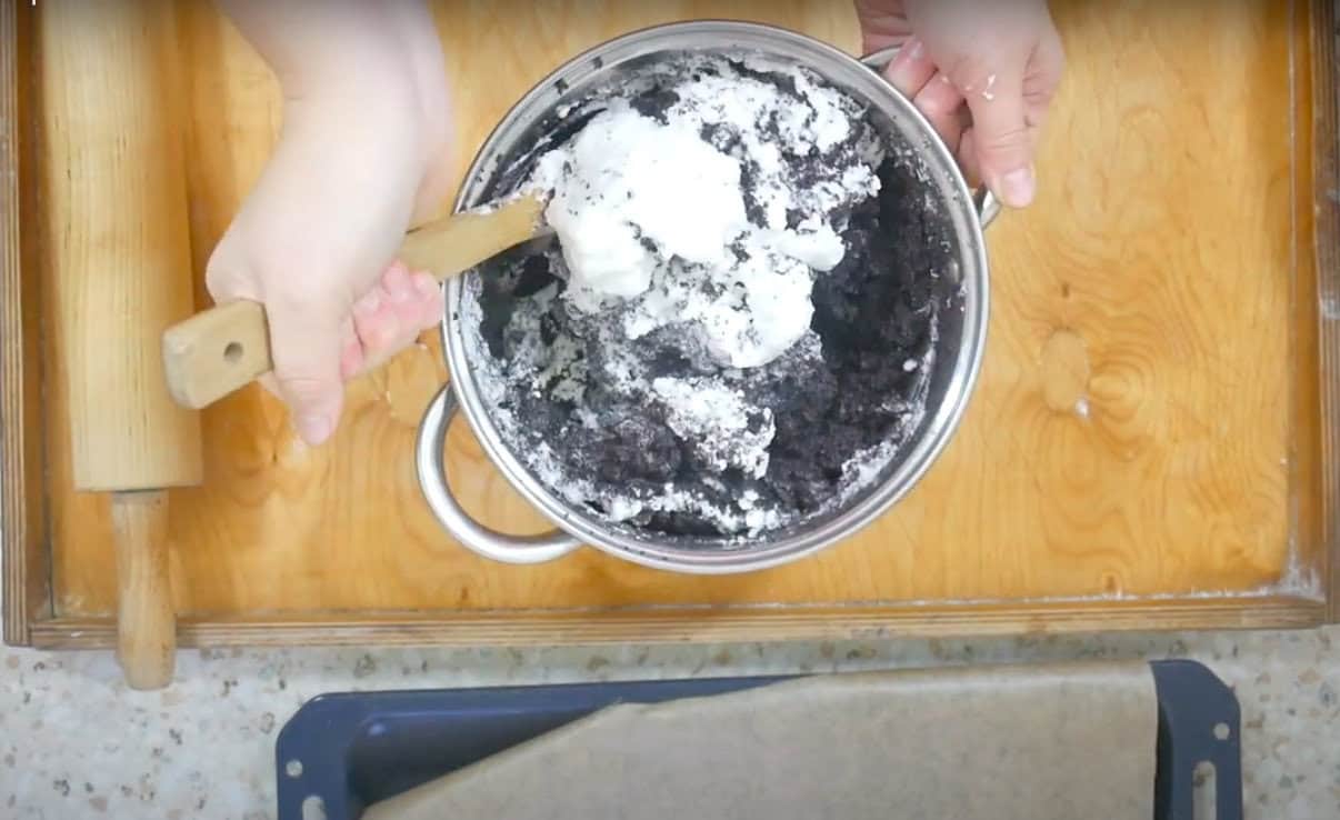 Overhead view of hands mixing white and black poppy seeds in a bowl with a wooden spoon on a kitchen counter for a Polish poppy seed roll recipe.