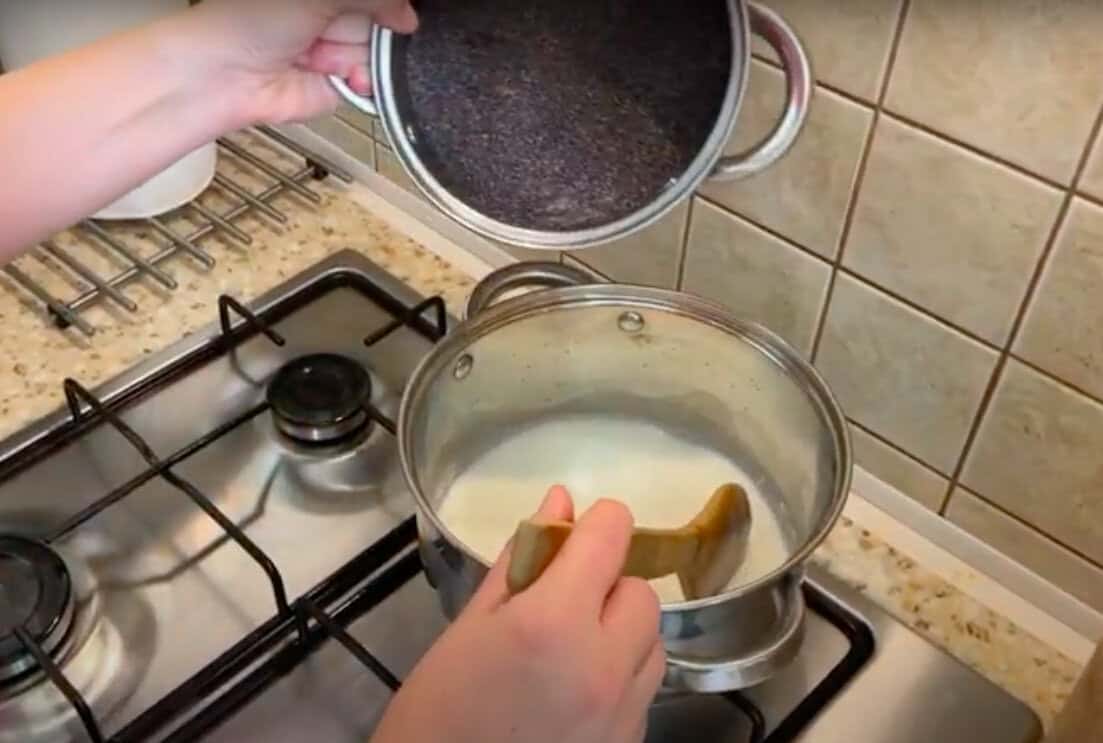 A person pours ground coffee from a metal pot into a saucepan of boiling water on a stove to make a Polish poppy seed roll recipe.
