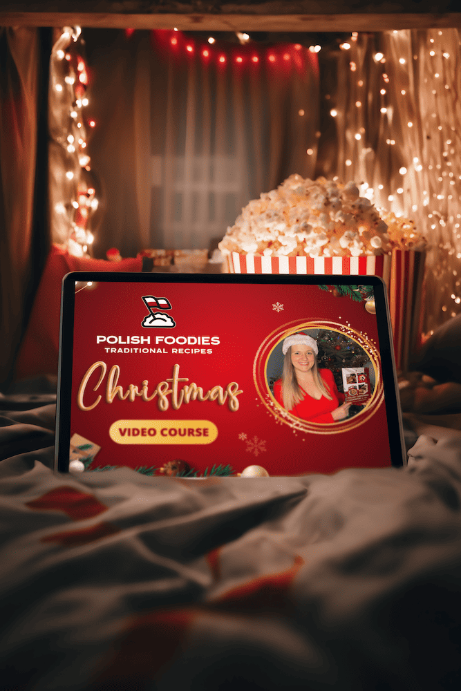 A tablet displaying a "Polish Foodies Traditional Recipes Christmas Video Course" sits on a bed surrounded by festive lights and a large bowl of popcorn, capturing the essence of Polish Christmas traditions.