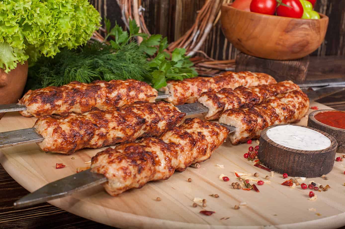 Grilled chicken kebabs on skewers with fresh herbs and sauces on a wooden board.