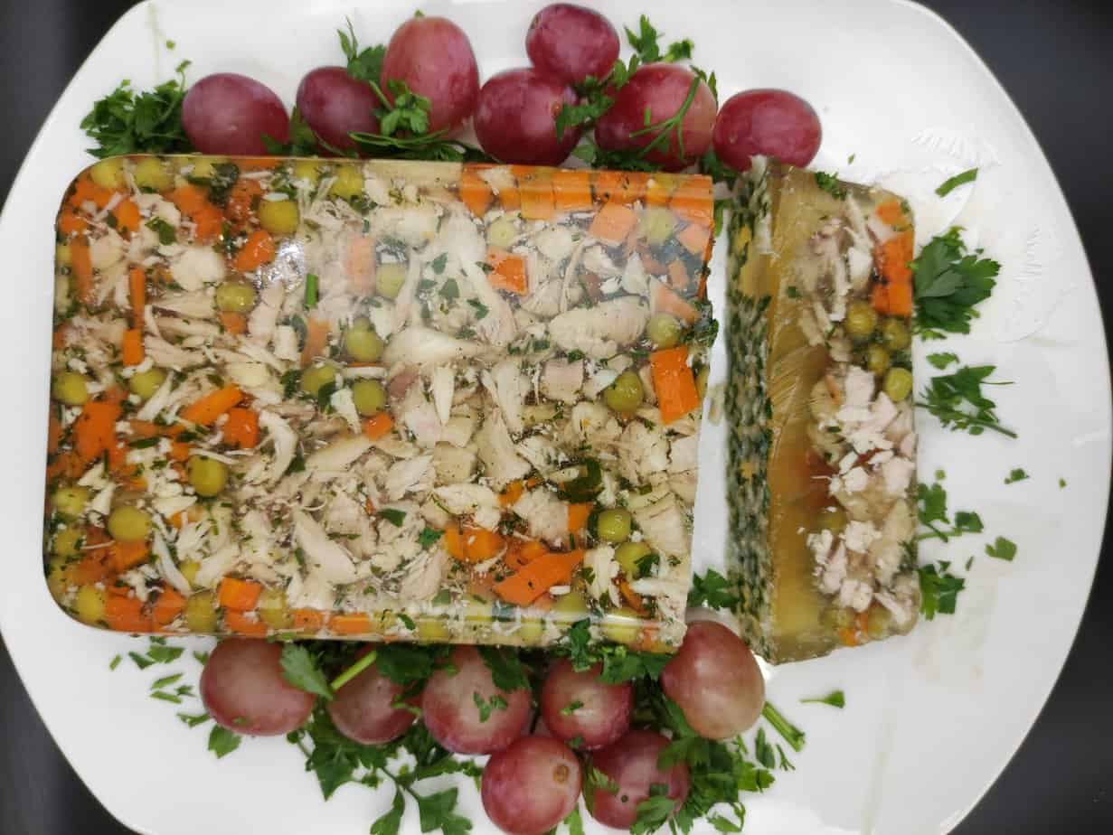 A rectangular Polish chicken and vegetable aspic garnished with parsley and surrounded by red grapes on a white plate.