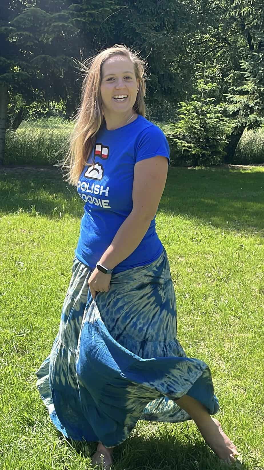 A smiling person wearing a blue t-shirt and a long, flowing skirt stands on green grass in a sunny outdoor setting, exuding warmth and openness that invites friendly contact.