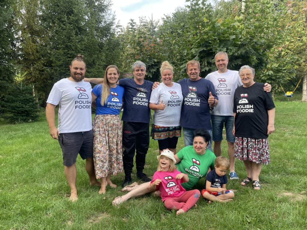 A group of ten people, including children, wearing "Polish Foodie Club" shirts, stand and sit together on a grassy area with trees in the background.