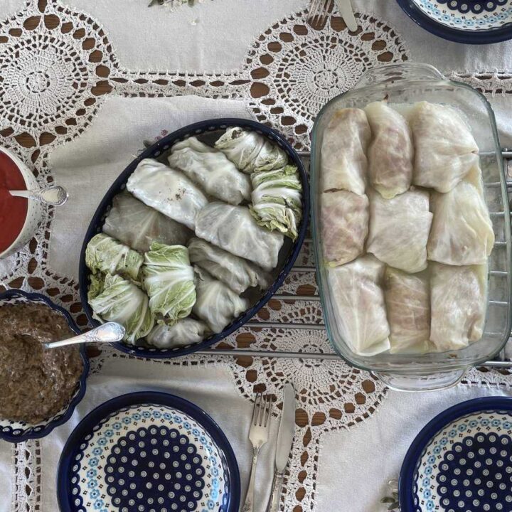 Two dishes of Gołąbki Z Ziemniakami rest on a lace tablecloth, accompanied by savory bowls of sauce and three place settings.