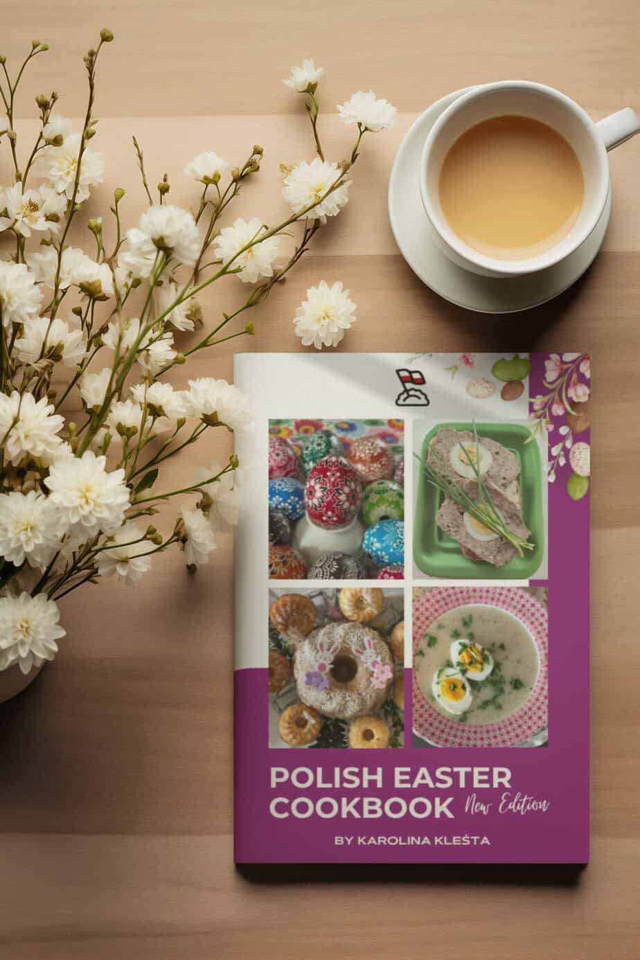 The "Polish Easter Cookbook" sits elegantly on the table beside a steaming cup of tea and a delicate bouquet of white flowers, its cover showcasing an array of festive dishes perfect for celebrating Easter traditions.