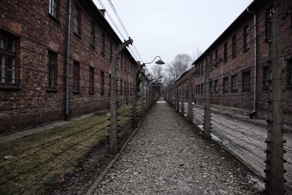Barbed wire fences separate two rows of brick buildings along a gravel path under a cloudy sky—one of the historic sites often included in the best day trips from Krakow.