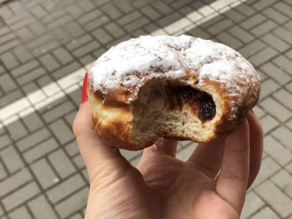 A hand holding a partially eaten jelly-filled doughnut topped with powdered sugar, set against a brick sidewalk—an irresistible taste of Wrocław food for those exploring the city’s sweet treats.