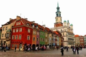 A town square with colorful historic buildings, a clock tower, and people walking on cobblestone streets under a cloudy sky—discover interesting Poznan as you soak in its charming atmosphere.