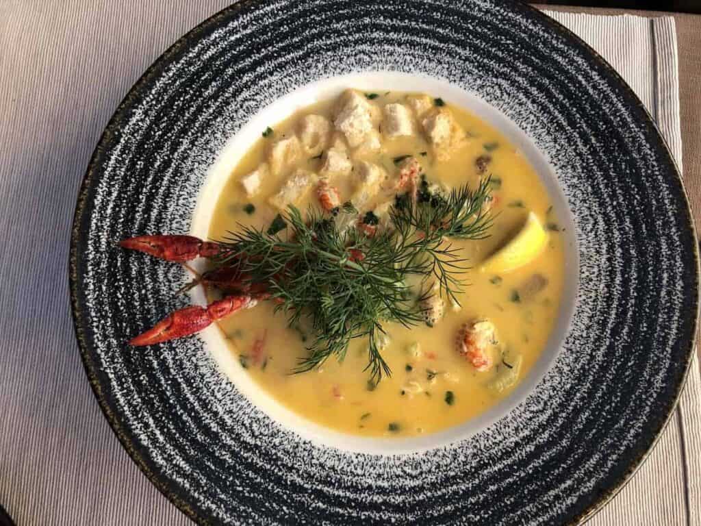 A bowl of creamy yellow seafood soup, garnished with fresh dill, croutons, lemon wedge, and crayfish claws on a speckled plate—a must-try for anyone searching where to eat authentic flavors in restaurants Wrocław offers.