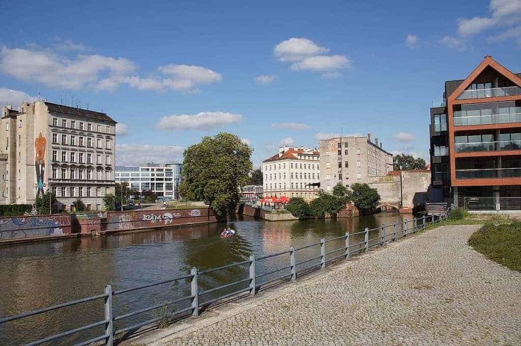 A riverside path with metal railing runs beside a calm river bordered by buildings, trees, and graffiti under a blue sky—perfect for discovering Wrocław restaurants or deciding where to eat Wrocław’s famous local food.