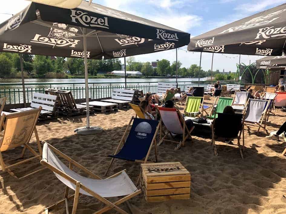 People relax on deck chairs under large Kozel umbrellas by the riverside, enjoying the sandy area surrounded by trees and water—a perfect spot if you’re looking for where to eat Wrocław style.