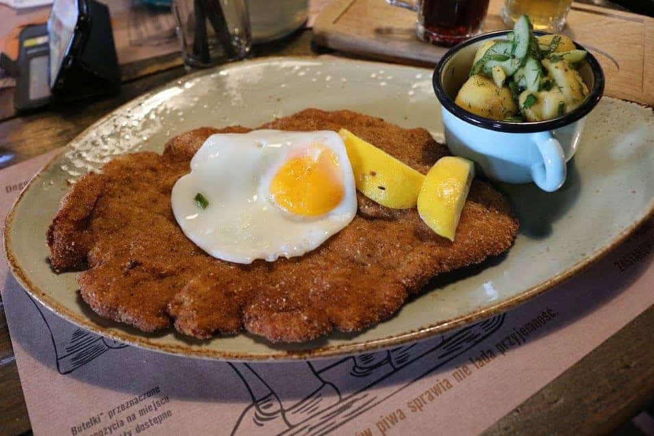 A plate with breaded schnitzel topped with a fried egg, lemon wedges on the side, and a small cup of boiled potatoes with herbs—a classic Wrocław food you’ll find in many top restaurants Wrocław has to offer.