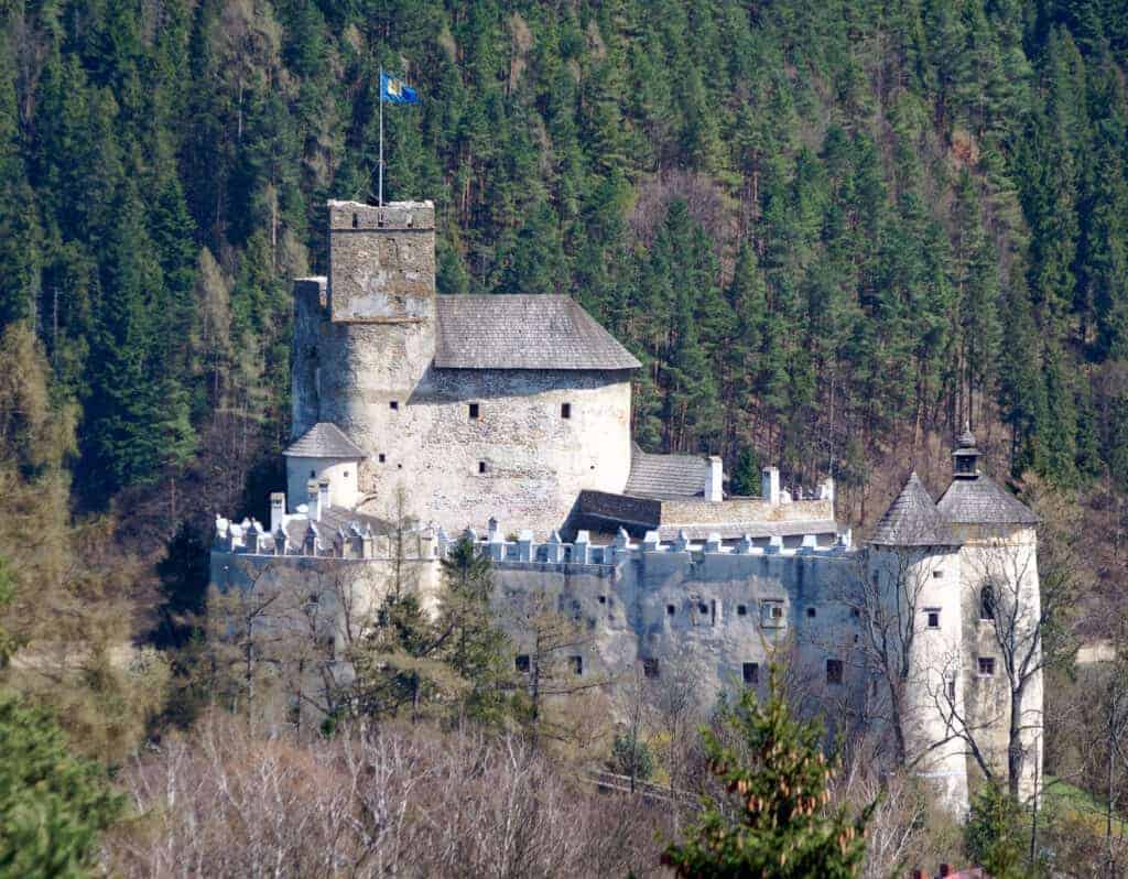 Discover one of the must-see castles in Poland: a stone castle with a central square tower and crenellated walls, surrounded by trees and forested hills in the background.
