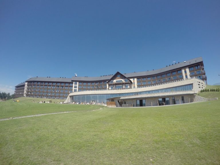 A large modern hotel with multiple stories and balconies is situated on a grassy lawn under a clear blue sky, offering an ideal retreat for exploring the Bieszczady Mountains in Poland—an insider’s guide to comfort and adventure.