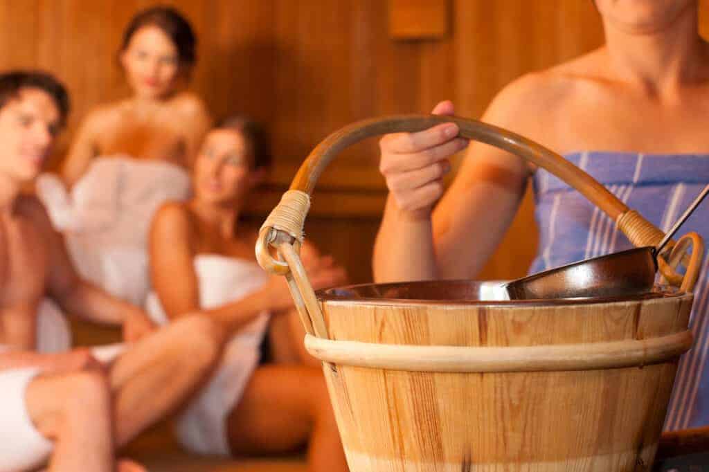 Three people in towels sit in a sauna at thermal baths Kraków, while another stands in the foreground holding a wooden bucket and ladle.