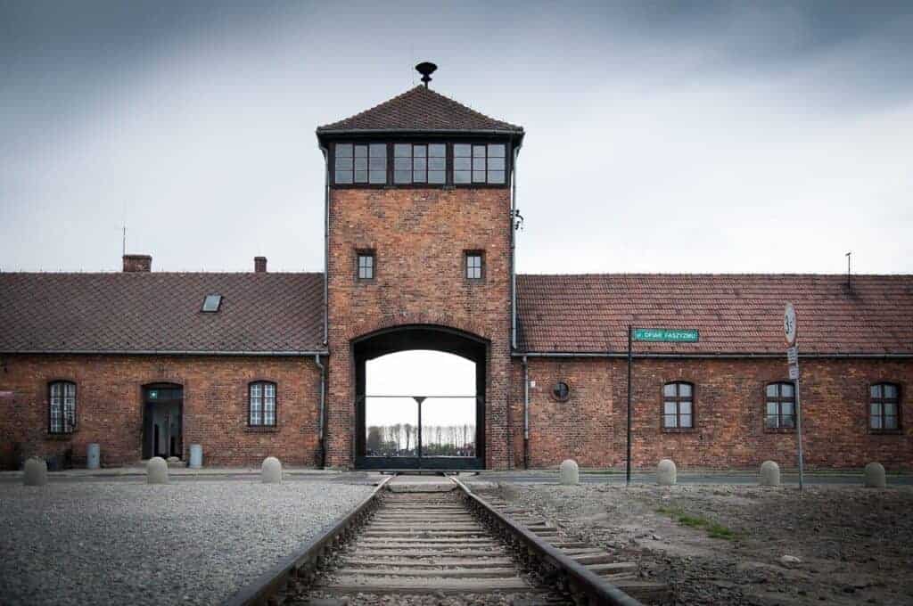 Brick entrance building of Auschwitz concentration camp with railway tracks leading inside, under a cloudy sky—a significant site to reflect on during travel in Poland.
