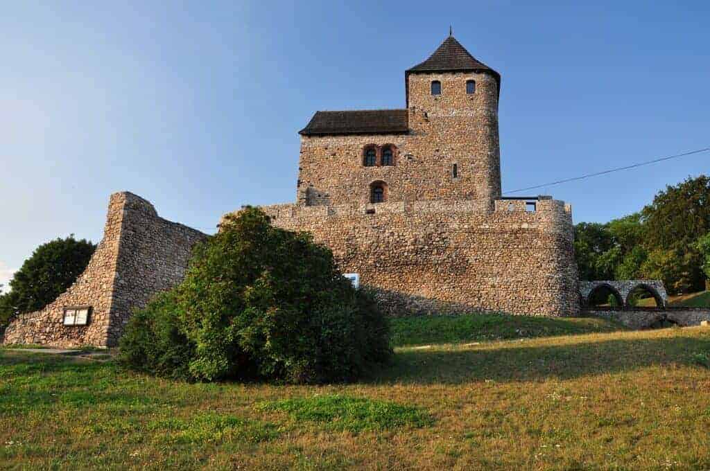 A stone medieval castle with a tall tower and surrounding walls sits on a grassy hill under a clear blue sky, making it one of the best places to visit in Slaskie, Poland.