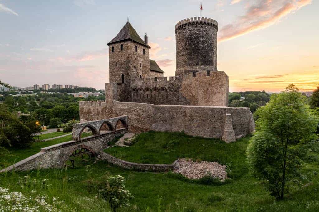 A medieval stone castle with a round tower and arched bridge sits on a grassy hill at sunset, surrounded by trees and distant buildings—a true gem among must-see castles in Poland.