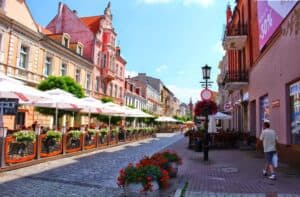 A cobblestone street lined with outdoor cafes, colorful historic buildings, flower pots, and a few pedestrians on a sunny day.