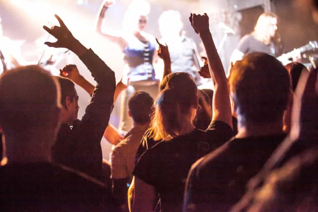 Crowd facing a brightly lit stage with raised hands at a live music concert; musicians and performers are visible in the background, capturing the vibrant energy of festivals in Poland.