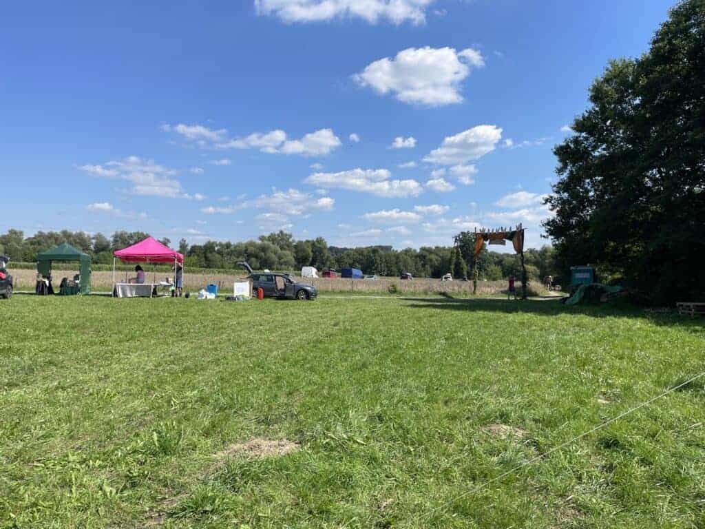 A grassy outdoor field with scattered tents, tables, a few people, some vehicles, and a wooden structure under a blue sky with clouds captures the vibrant scene found at some of the Best Festivals Poland has to offer.