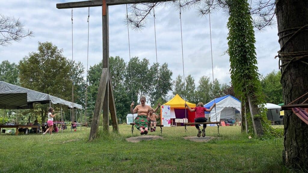 Two people are sitting on swings in a grassy outdoor area at one of the best festivals in Poland, with tents and trees in the background. Other people are seen walking and standing nearby, enjoying the lively atmosphere of Polish festivals.