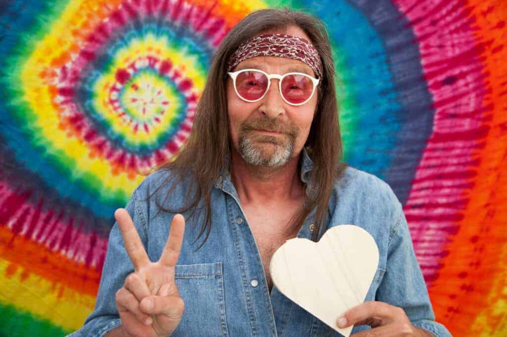 Man with long hair and glasses, wearing a headband and denim shirt, holds a white heart and makes a peace sign in front of a colorful tie-dye background, capturing the vibrant spirit of Polish festivals.