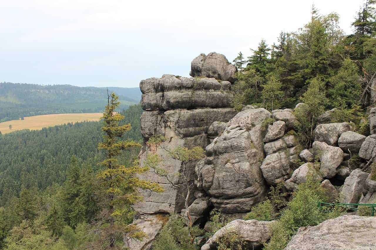 Large rock formations with layered stone surfaces rise above a forested landscape, with trees and distant hills visible under a cloudy sky—an iconic view when you visit Dolnośląskie, Poland.