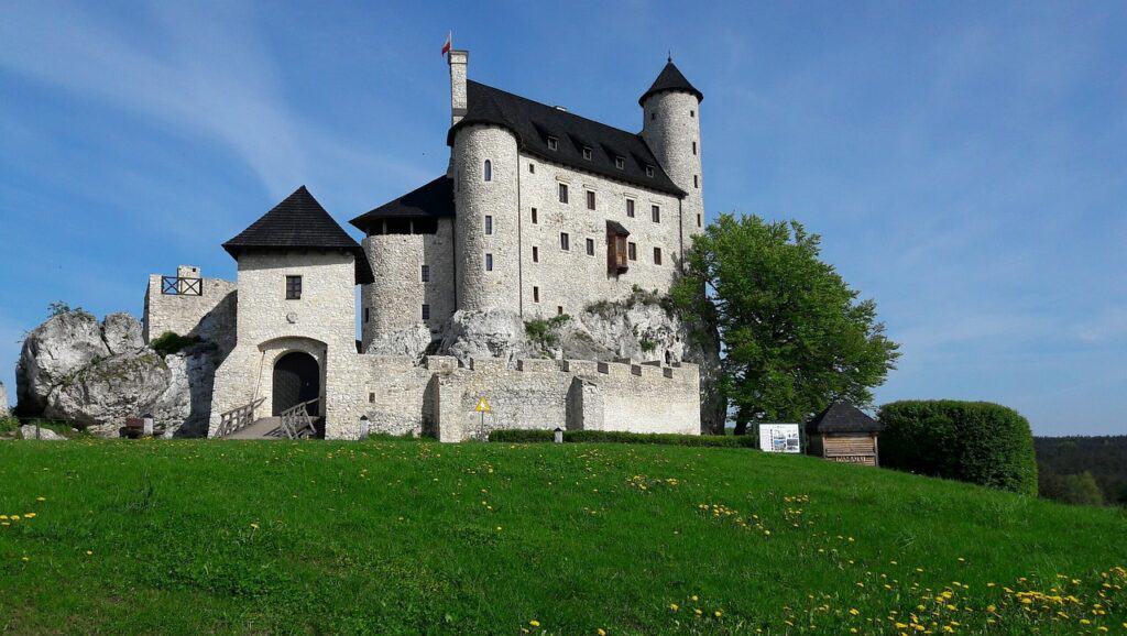 A stone castle with round towers and peaked roofs stands on a grassy hill under a blue sky, surrounded by a stone wall and some trees—one of the best places to visit in Slaskie, Poland for history and travel lovers.