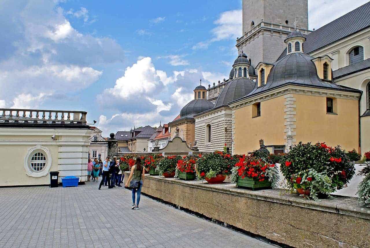 People walk along a paved terrace lined with red flowers in pots, adjacent to historic buildings with domed roofs under a partly cloudy sky—an inviting scene among the best places to visit in Slaskie, Poland for travel enthusiasts.