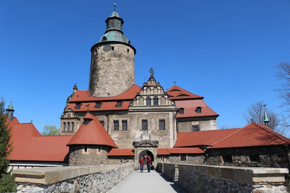 A stone castle with a cylindrical tower and red-tiled roofs is viewed from a walkway under a clear blue sky, showcasing why Dolnośląskie, Poland is among the best places to visit.