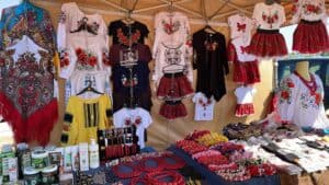 A market stall displays traditional embroidered clothing, including blouses and skirts, along with jewelry, scarves, and cosmetic products on tables.