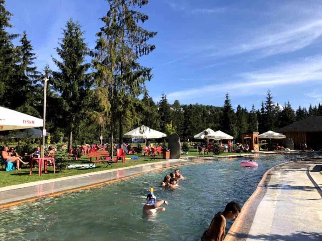 People are swimming and relaxing in an outdoor pool surrounded by pine trees, while others unwind under umbrellas on red chairs nearby—perfect for those seeking Zakopane thermal baths or looking for Zakopane tips.