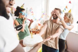 Group of adults at a birthday party; one person holds a cake, another is blindfolded, others wear party hats and glasses. Decorations and a foosball table set the scene as they celebrate Name Day in Poland, honoring beloved Polish traditions.