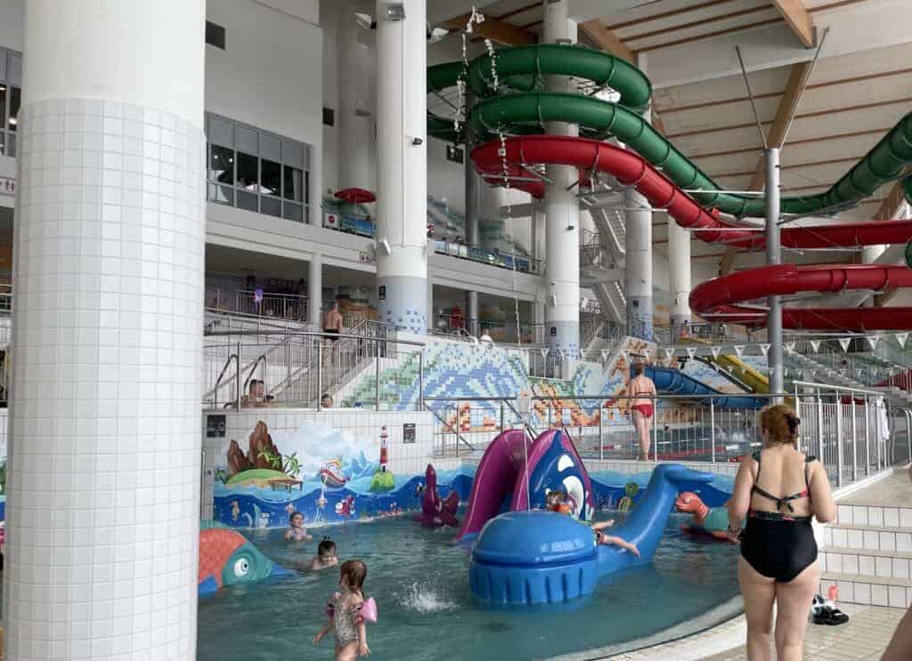Indoor water park with children playing in a shallow pool featuring aquatic-themed play structures; large water slides and people visible in the background at Zakopane thermal spas.