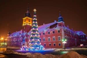 A large Christmas tree with colorful lights and presents stands in front of a historic building at night, with snow on the ground and building rooftops.