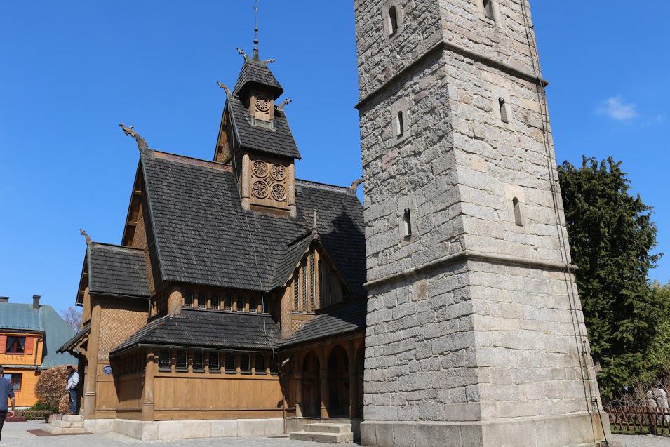 A wooden stave church with ornate carvings stands beside a tall stone bell tower under a clear blue sky—one of the best places in Dolnośląskie Poland to explore history and architecture.