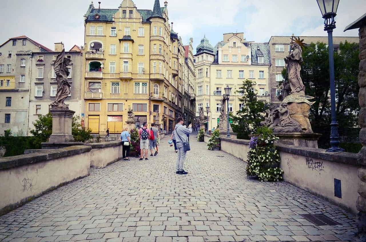 People walk across a cobblestone bridge with statues and flower planters, surrounded by historic European buildings under a partly cloudy sky—one of Dolnośląskie’s best places to visit for unforgettable Poland travel experiences.