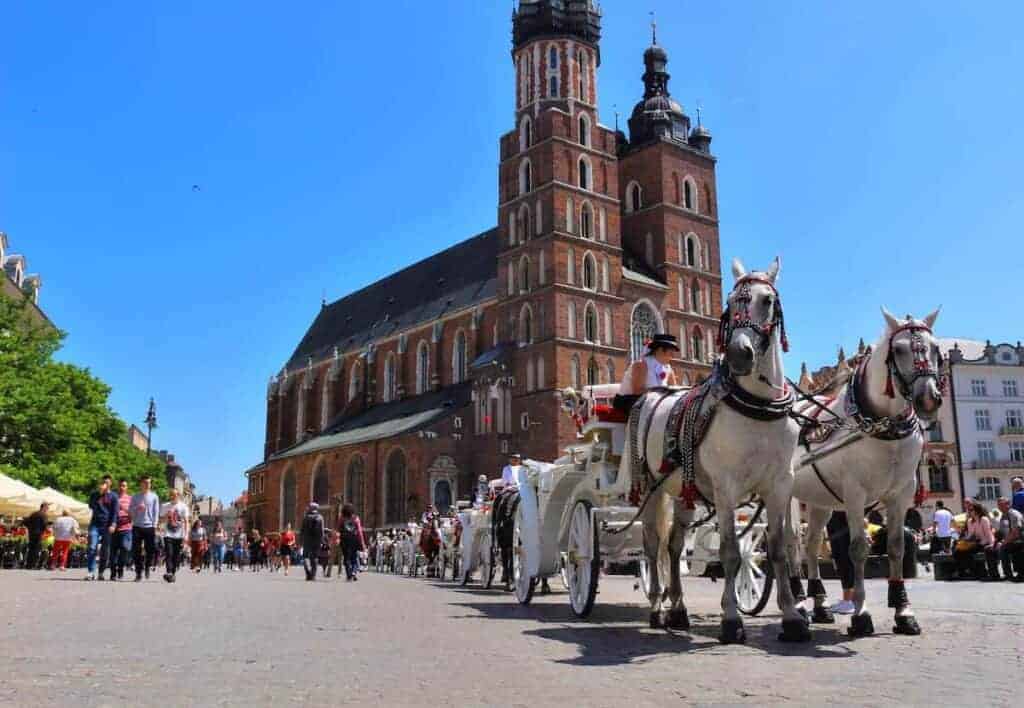 A white horse-drawn carriage passes in front of a large brick church with twin towers on a sunny day, as people stroll nearby—capturing the charm of a Poland city often counted among the top cities Poland has to offer.