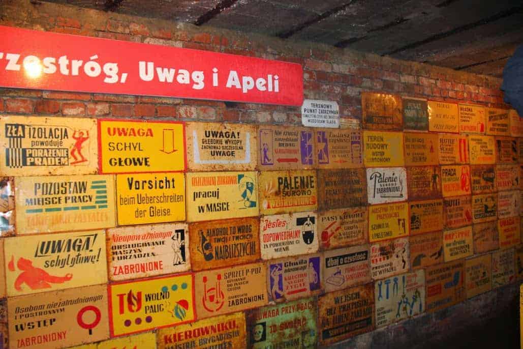 A brick wall covered with numerous vintage warning and prohibition signs in Polish and German, displayed under a red banner with white text—a unique sight for Poland travel enthusiasts exploring Dolnośląskie tourism.