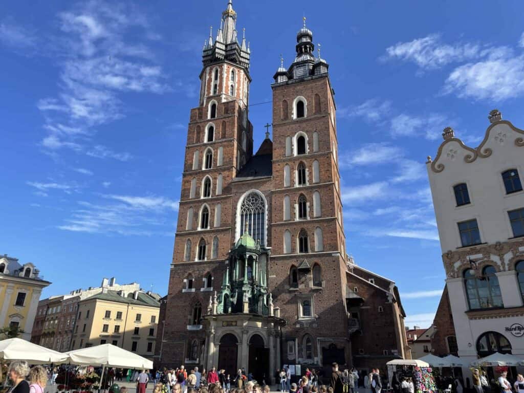 A large brick church with two tall towers and a central entrance stands in a busy square under a blue sky, surrounded by historic buildings and people—one of the top things to see in Poland on any 5 days itinerary.