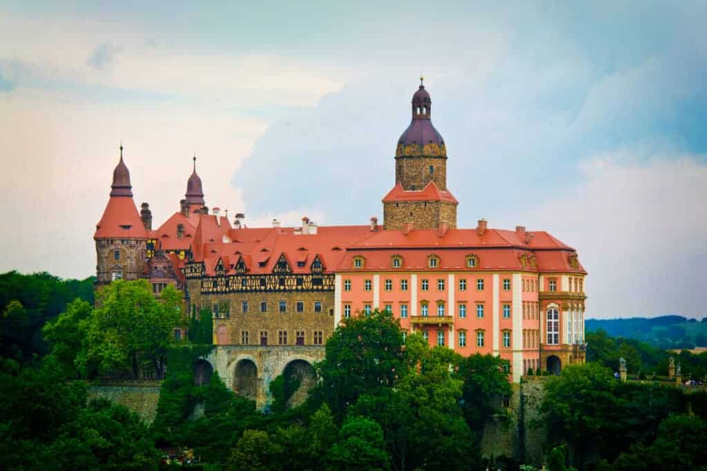 A large castle with red roofs and multiple towers sits atop a hill surrounded by trees under a cloudy sky—one of the must-see castles that stands out among Poland attractions.