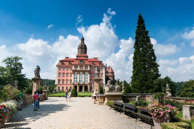 People walk along a stone pathway leading to a large, historic pink castle—one of the best places to visit in Dolnośląskie, Poland—surrounded by statues, benches, plants, and a tall evergreen tree under a blue sky.